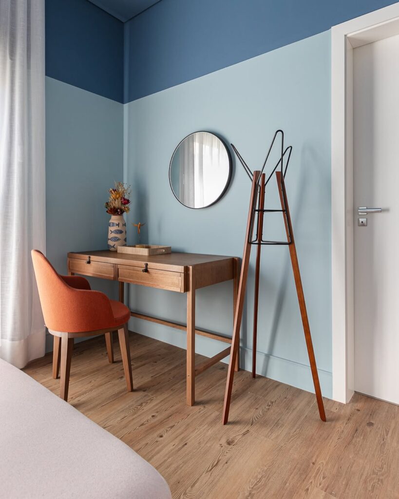 Quarto com parede em dois tons de azul, espelho redondo e mancebo ao lado da escrivaninha