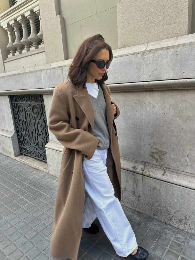 Mulher jovem, magra e de pele clara. Ela tem cabelos curtos e usa óculos de sol e está caminhando na rua. Ela usa sobretudo, suéter, calça jeans branca e sapatilha