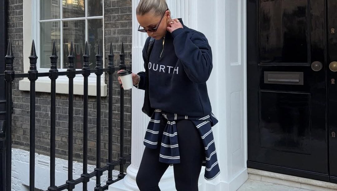Mulher jovem de pele clara com cabelo preso em um coque. Ela usa moletom, calça legging, tênis e tem suéter amarrado na cintura