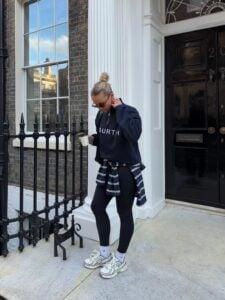 Mulher jovem de pele clara com cabelo preso em um coque. Ela usa moletom, calça legging, tênis e tem suéter amarrado na cintura