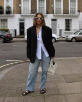 Mulher jovem, de pele clara e com cabelo curto liso. Ela usa blazer oversized preto, camisa branca, calça jeans e rasteira preta.