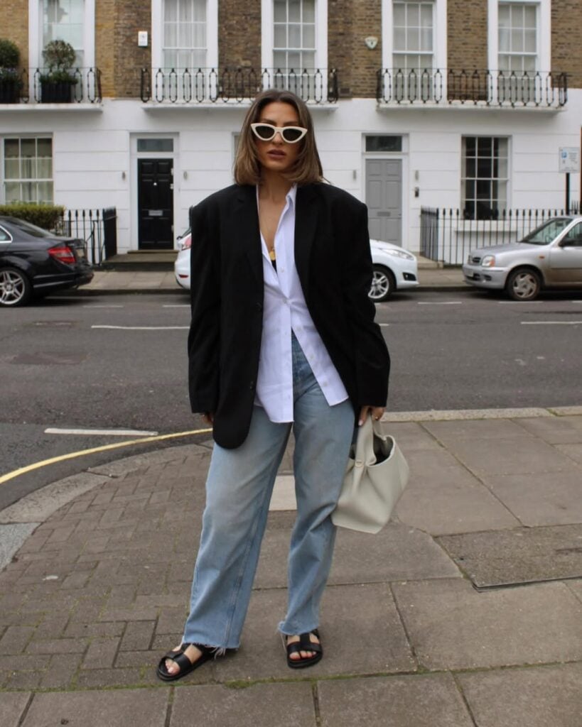 Mulher jovem, de pele clara e com cabelo curto liso. Ela usa blazer oversized preto, camisa branca, calça jeans e rasteira preta.