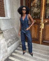 Mulher jovem, magra e de pele negra. Ela usa colete jeans, calça jeans e sapato de bico fino