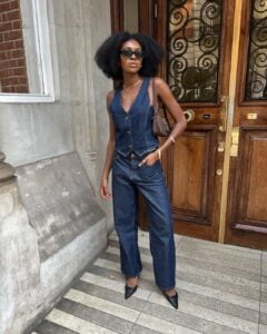 Mulher jovem, magra e de pele negra. Ela usa colete jeans, calça jeans e sapato de bico fino