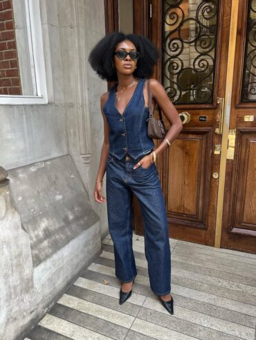 Mulher jovem, magra e de pele negra. Ela usa colete jeans, calça jeans e sapato de bico fino