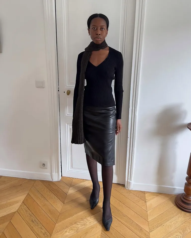 Mulher jovem, magra e de pele negra. Ela usa blusa de manga longa, saia lápis, meia-caçlça e sapato de bico fino