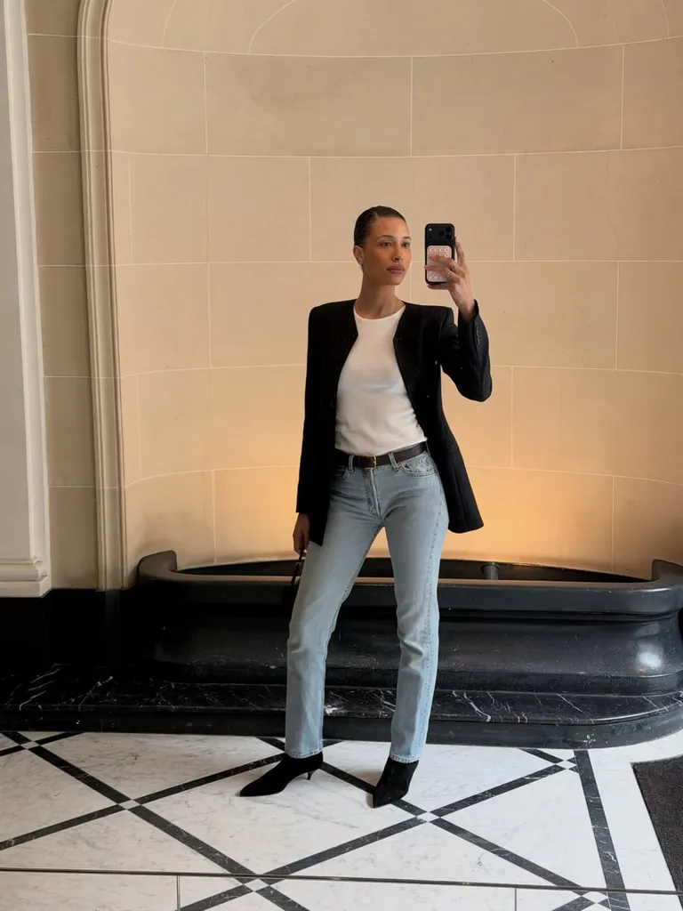 Mulher jovem, magra e de pele clara. Ela tem cabelo castanho preso em um coque e usa blazer, blusa básica, calça jeans, cinto e bota com salto alto