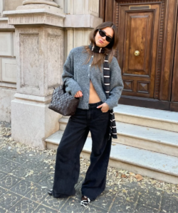 Mulher jovem, magra e de pele clara com cabelo castanho claro. Ela usa cardigan, calça jeans e sapatilha
