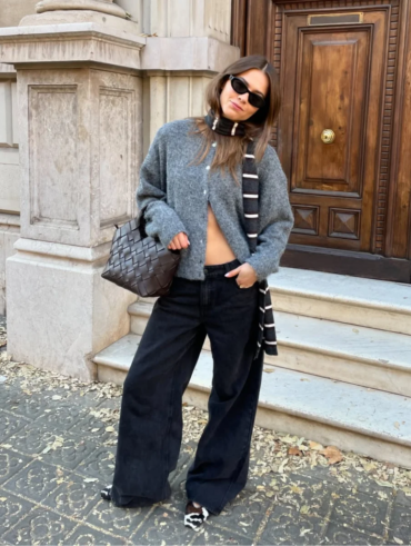 Mulher jovem, magra e de pele clara com cabelo castanho claro. Ela usa cardigan, calça jeans e sapatilha