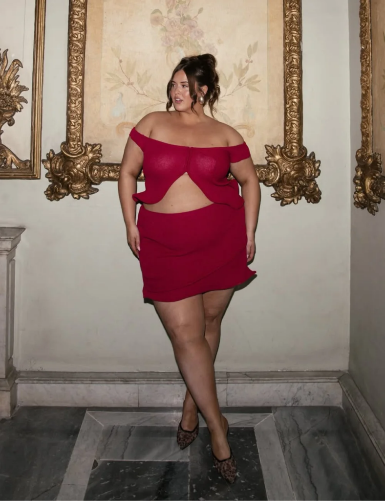Mulher jovem, de pele clara e cabelo castanho preso em coque. Ela está posando para uma foto na frente de um quadro grande. Ela veste conjunto de top com saia curta na cor vermelha e sapatilha de bico fina estampada