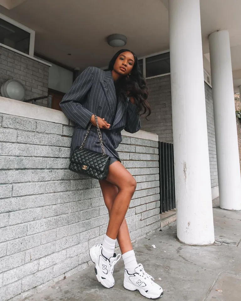 Mulher jovem, magra, de pele negra e com cabelo escuro ondulado. Ela usa blazer, bermuda, tênis New Balance e meias brancas