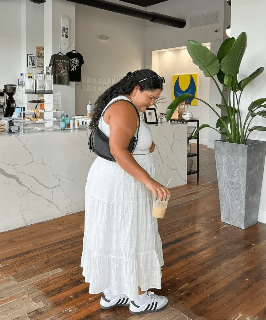Mulher jovem e gorda de pele clara e cabelo escuro médio. Ela usa regata branca, saia longa branca, tênis Adidas Samba e bolsa meia lua