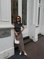 Mulher jovem, magra e de pele clara com cabelos lisos e castanhos. Ela usa blusa cinza, saia de alfaiataria longa marrom claro e scarpin branco