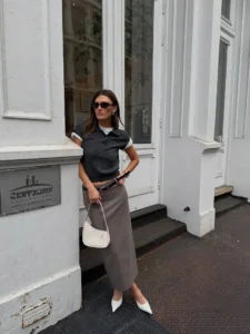 Mulher jovem, magra e de pele clara com cabelos lisos e castanhos. Ela usa blusa cinza, saia de alfaiataria longa marrom claro e scarpin branco