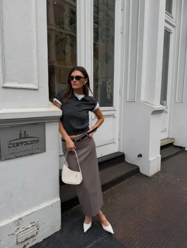 Mulher jovem, magra e de pele clara com cabelos lisos e castanhos. Ela usa blusa cinza, saia de alfaiataria longa marrom claro e scarpin branco
