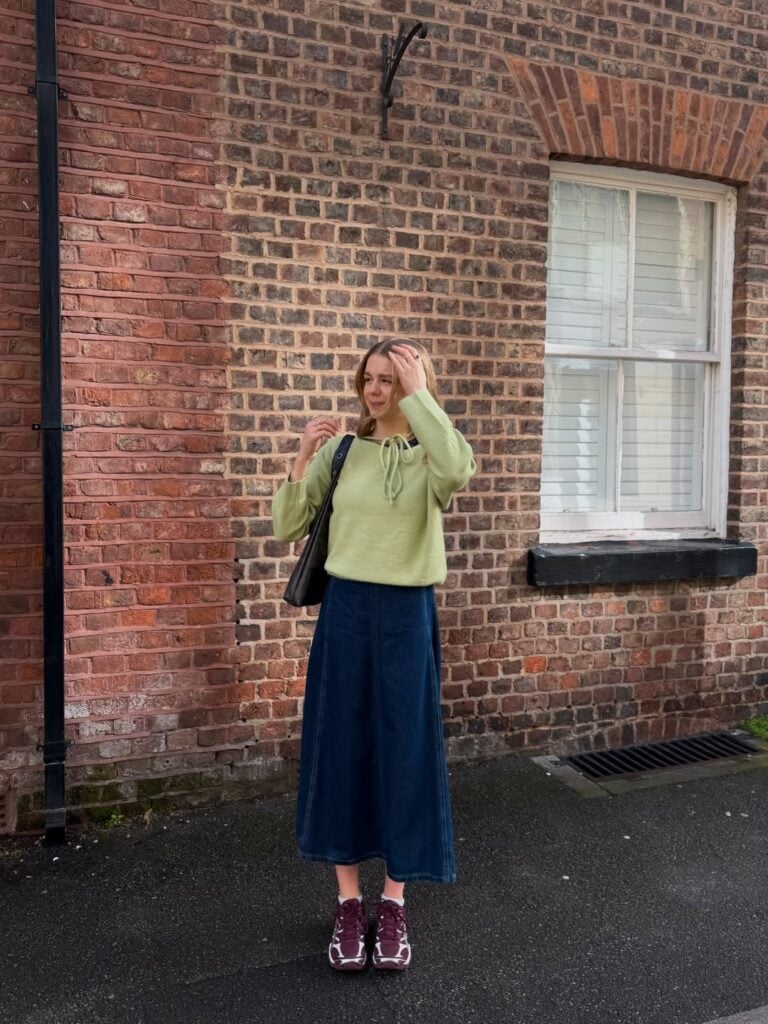 Mulher jovem, magra e de pele clara. Ela usa look com saia jeans, suéter e tênis