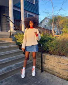 Mulher jovem, magra e de pele negra com cabelo cacheado curto. Ela usa suéter, saia jeanis curta e tênis