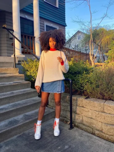 Mulher jovem, magra e de pele negra com cabelo cacheado curto. Ela usa suéter, saia jeanis curta e tênis