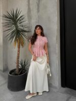 Mulher jovem, magra e de pele clara com cabelo castanho ondulado. Ela usa blusa rosa, saia longa branca e sapatilha tabi branca