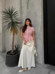 Mulher jovem, magra e de pele clara com cabelo castanho ondulado. Ela usa blusa rosa, saia longa branca e sapatilha tabi branca