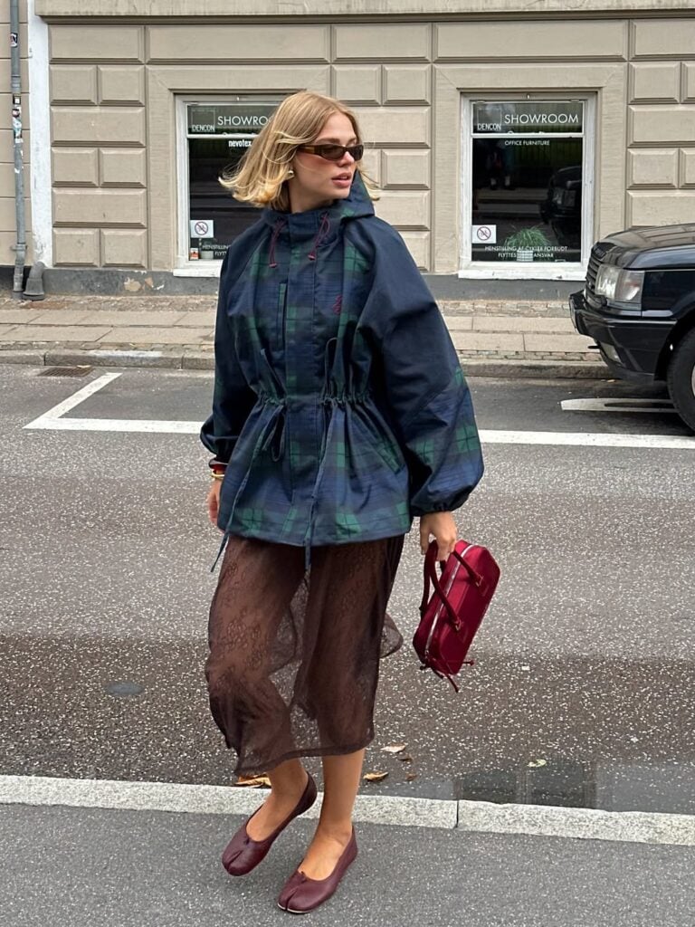 Mulher jovem, magra, com cabelo curto claro e de pele clara. Ela usa jaqueta, saia e sapatilha tabi