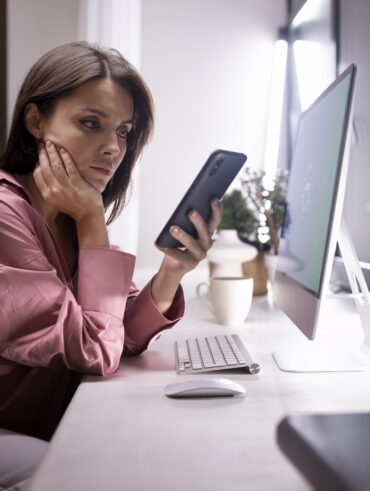 Mulher com cabelo escuro e camisa rosa em frente ao computador, com celular na mão