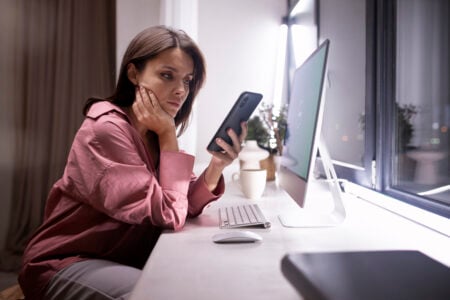 Mulher com cabelo escuro e camisa rosa em frente ao computador, com celular na mão
