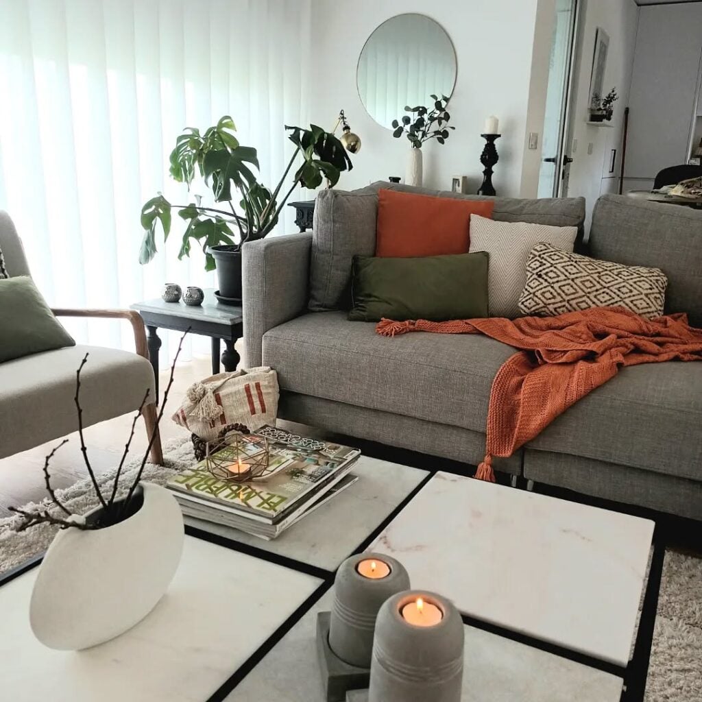 Sala de estar com sofá cinza e almofadas em tons terrosos, mesa de centro com revistas, vaso de cerâmica e velas