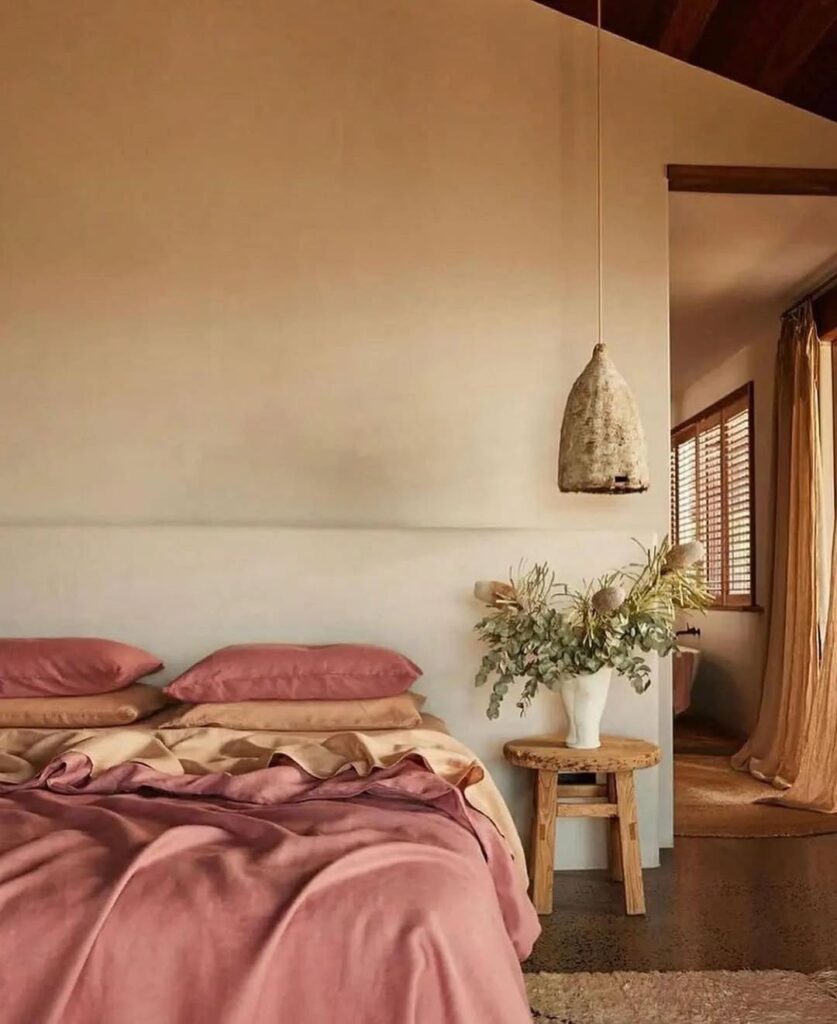 Quarto com lençóis e fronhas rosados, parede branca, banquinho ao lado da cama com planta