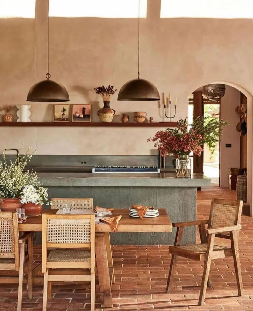 Cozinha com mesa e cadeiras de madeira, ilha em tom acinzentado, pendentes de metal e vasos de cerâmica com flores