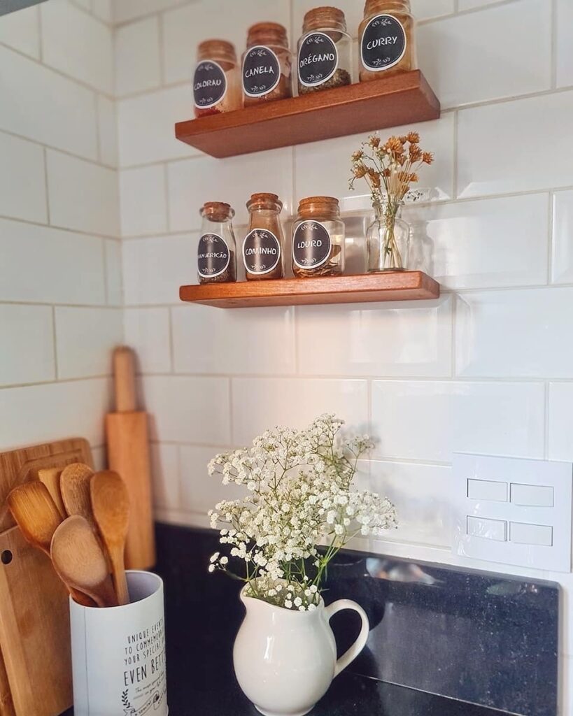 Cozinha com revestimento de tijolinhos brancos, prateleiras flutuantes de madeira, bancada escura com utensílios de bambu e jarrinha branca com flores
