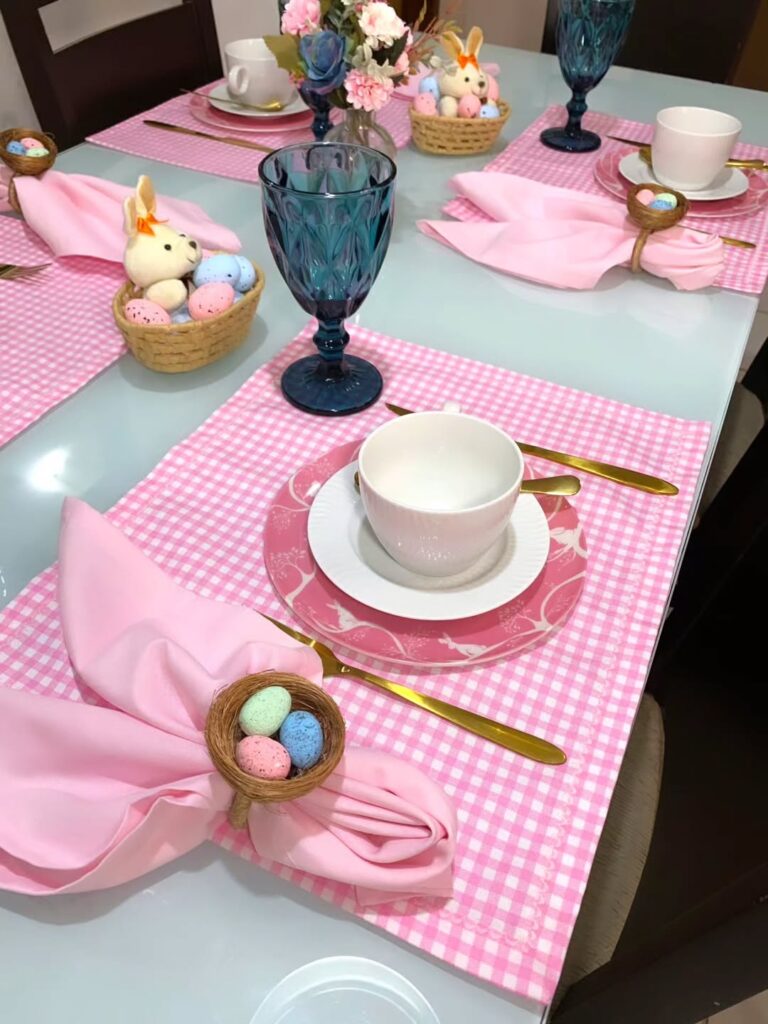 Mesa posta de páscoa com jogo americano xadrez em rosa e branco, guardanapo dobrado em formato de orelhas de coelho e taças azuis