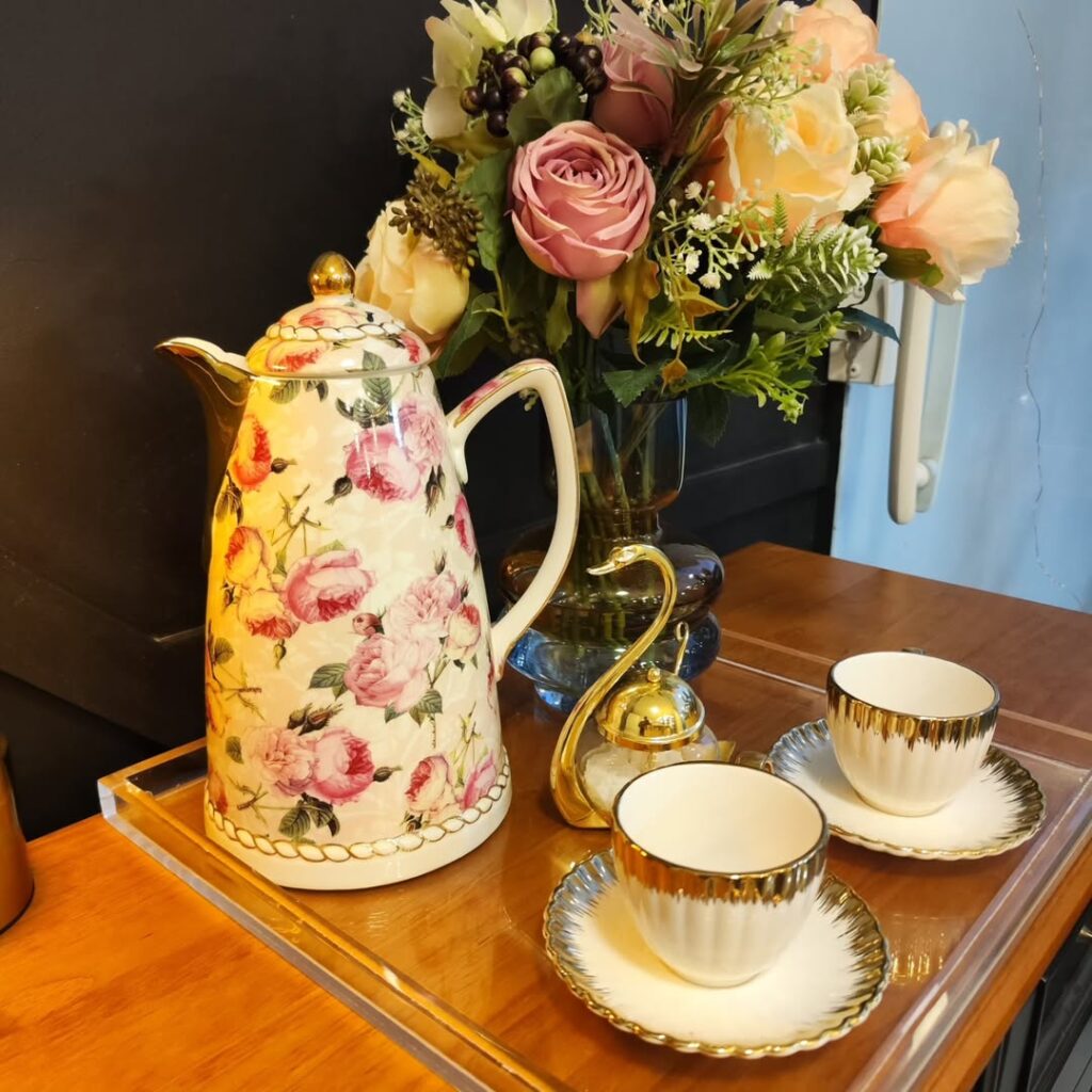 Bandeja de vidro com bule de porcelana e estampa floral, vaso de flores e xícaras claras com bordas douradas