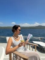Mulher de pele clara, com regata branca, cabelo preso. Ela está lendo um livro em um barco