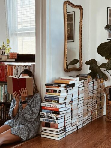 Mulher de pele clara sentada, lendo, com livros atrás