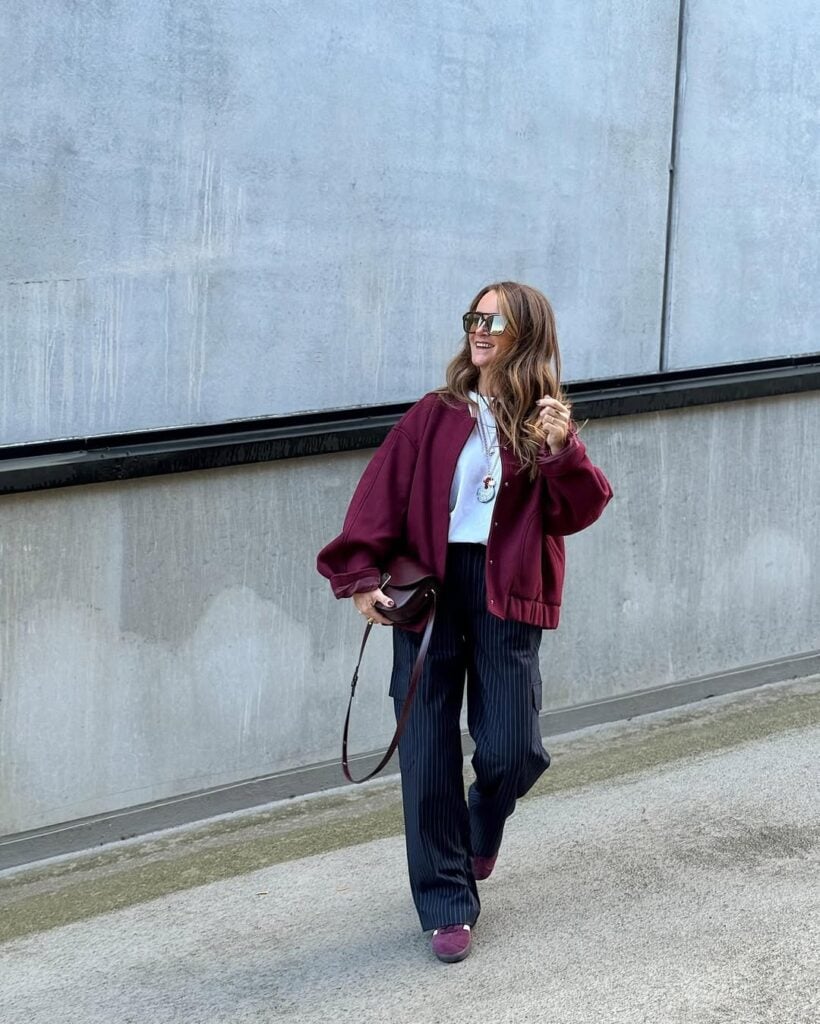 Mulher jovem de pele clara e cabelo loiro mel. Ela veste jaqueta bomber bordô, blusa branca, calça risca de giz e tênis casual bordô