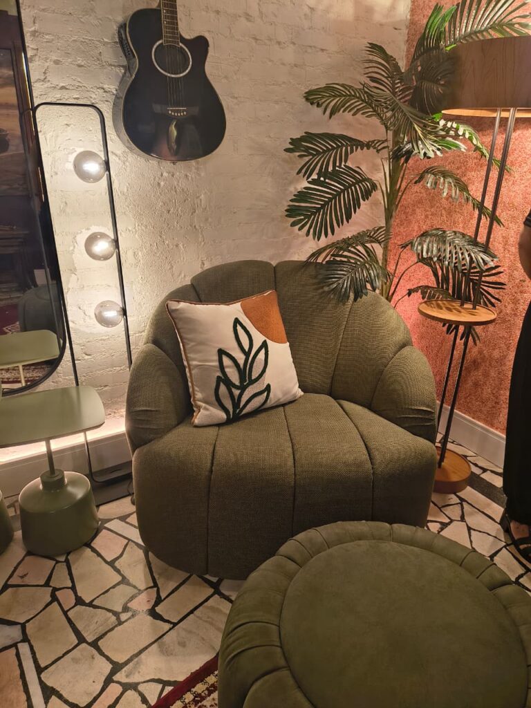 Cantinho com poltrona e peseira estofadas verde-musgo, com planta, luz baixa e violão na parede