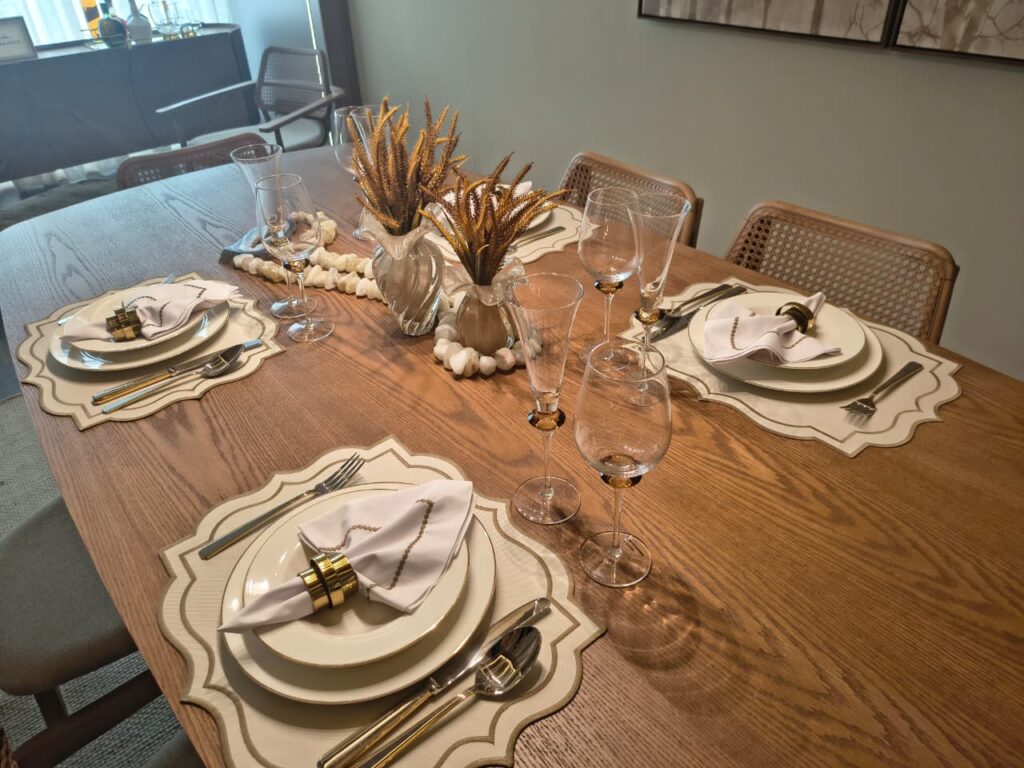 Mesa de jantar de madeira com jogo americano e louças claras, talheres com cabos dourados e arranjos secos