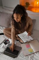 Mulher sentada sobre a cama com notebook, calculadora e cadernos