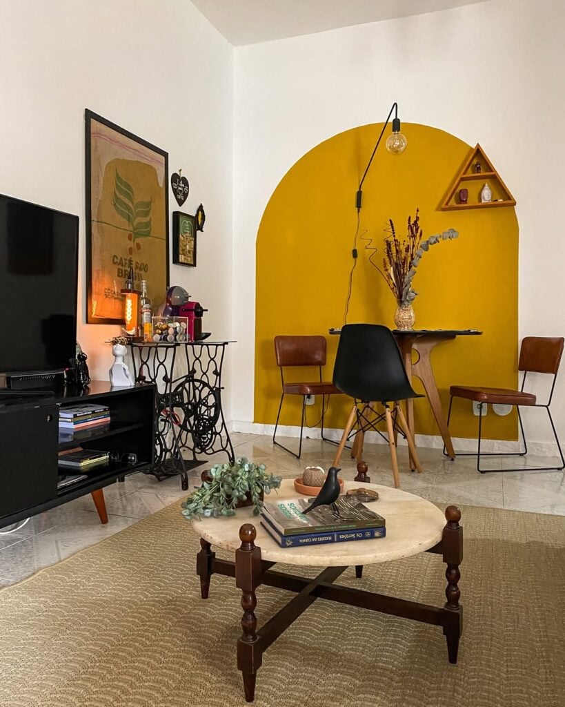 Sala de estar com paredes brancas e pintura orgânica em tom de mostarda onde fica a mesa e cadeiras