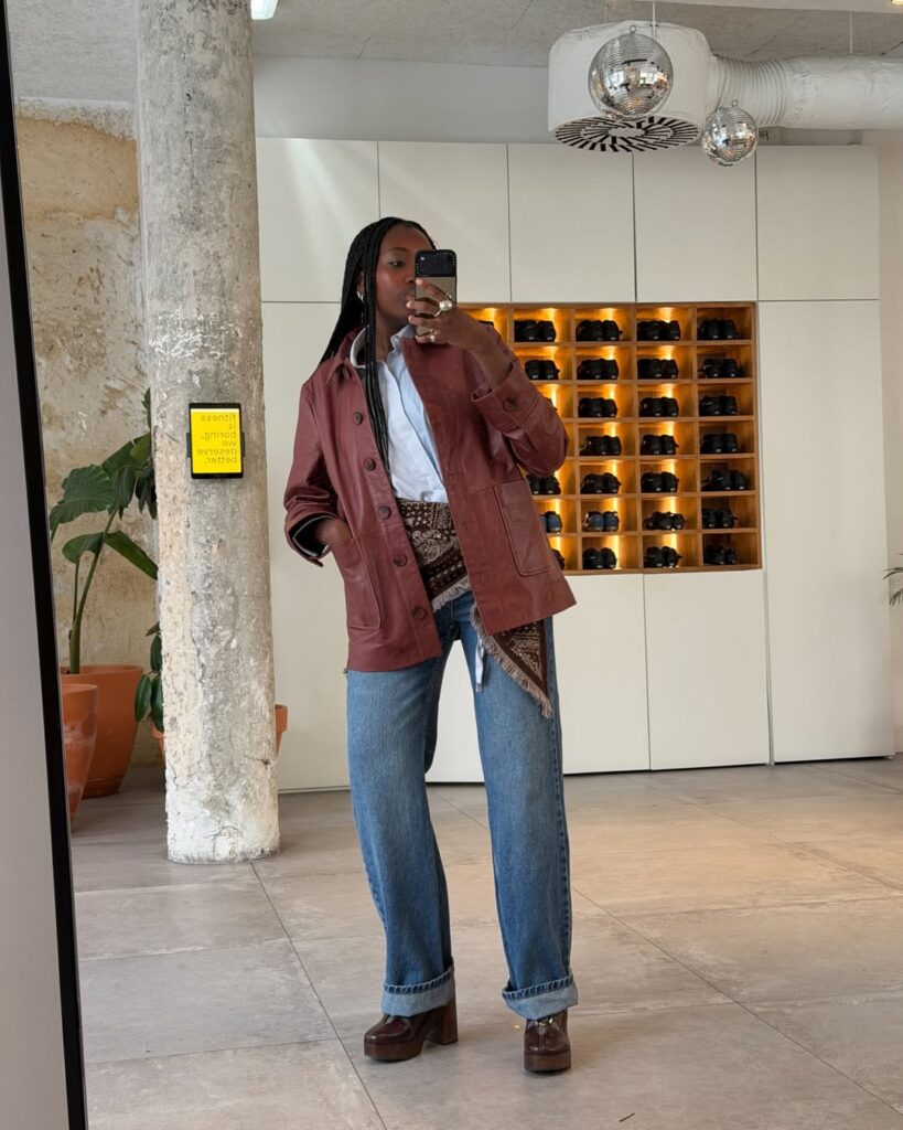 Mulher jovem de pele negra. Ela usa look de outono com jaqueta, camisa, calça jeans e bota com salto alto,