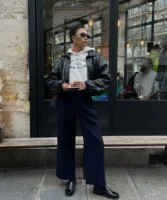 Mulher jovem e magra. Ela usa moletom, jaqueta bomber preta por cima, calça jeans azul escuro e mocassim preto.