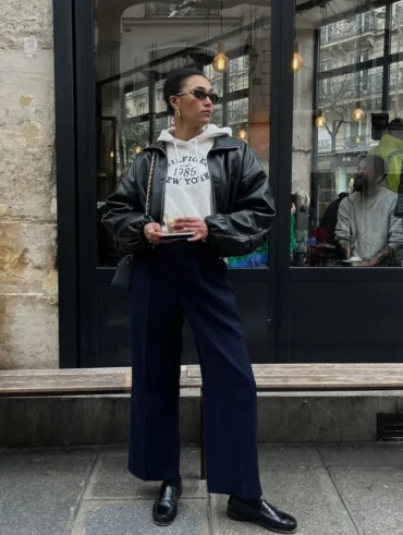 Mulher jovem e magra. Ela usa moletom, jaqueta bomber preta por cima, calça jeans azul escuro e mocassim preto.