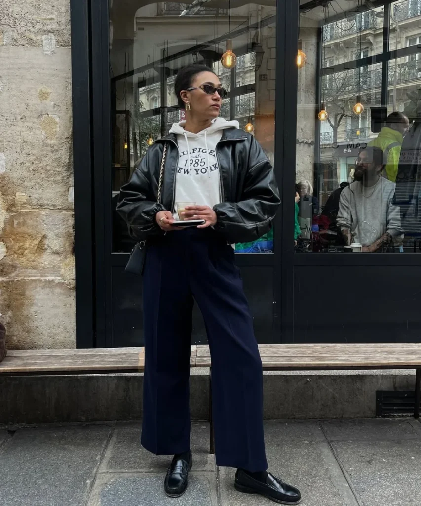 Mulher jovem e magra. Ela usa moletom, jaqueta bomber preta por cima, calça jeans azul escuro e mocassim preto.