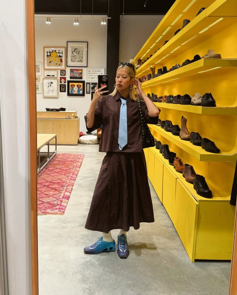 Mulher jovem, magra e de pele clara. Ela usa look marrom com gravata azul e tênis esportivo