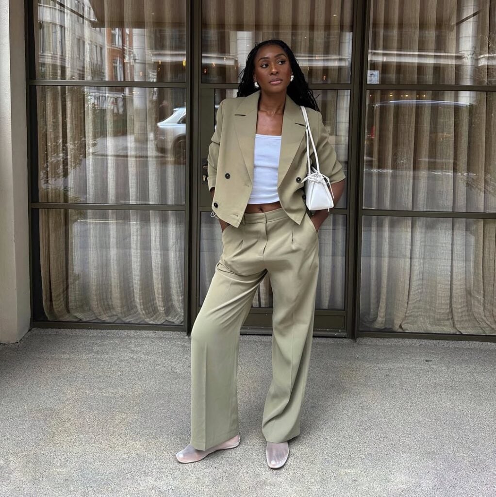 Mulher jovem, magra e de pele negra com cabelos longos. Ela usa blazer cropped, calça de alfaiataria, blusa branca e sapatilha telada