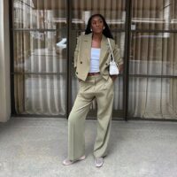 Mulher jovem, magra e de pele negra com cabelos longos. Ela usa blazer cropped, calça de alfaiataria, blusa branca e sapatilha telada