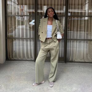 Mulher jovem, magra e de pele negra com cabelos longos. Ela usa blazer cropped, calça de alfaiataria, blusa branca e sapatilha telada