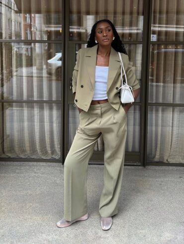 Mulher jovem, magra e de pele negra com cabelos longos. Ela usa blazer cropped, calça de alfaiataria, blusa branca e sapatilha telada