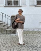 Mulher jovem de pele clara. Ela usa suéter marrom, saia acetinada off-white e tênis marrom claro